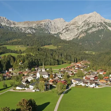 A picturesque mountain landscape with a small village amidst green meadows. In the background, high mountains rise under a clear blue sky.