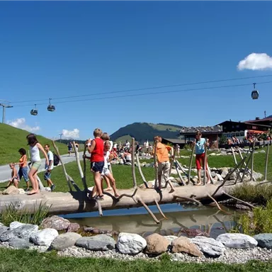 Children are playing on a wooden walkway over a small pond. In the background, mountains and a cable car can be seen.