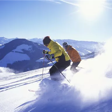 Two skiers are skiing down the snow-covered slope. The landscape is surrounded by mountains and clear, sunny sky.
