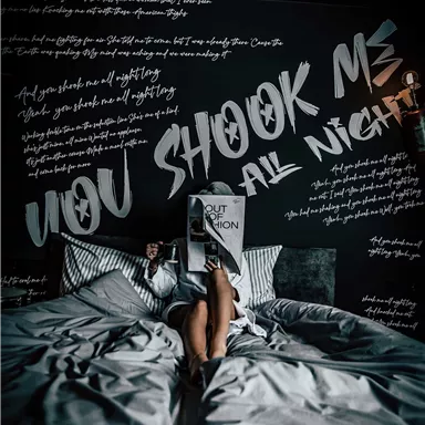 A cozy bedroom with a large bed and a black-painted mural. A person is sitting in the bed holding a book in front of their face.