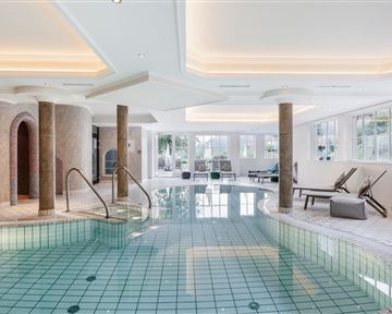 A luxurious indoor pool with a smooth, green tiled floor. The environment is bright and modern, with large windows and comfortable loungers.
