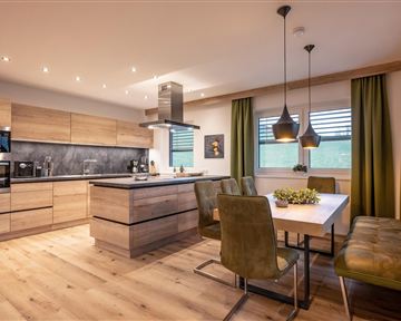 Eine moderne Küche mit Holzregalen und einem großen Esstisch. Die offene Atmosphäre wird durch die eleganten Lampen und die grünen Vorhänge unterstrichen.