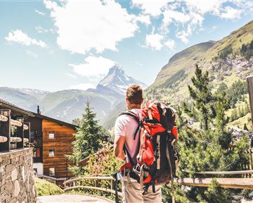 Ein Wanderer mit einem Rucksack steht vor einer beeindruckenden Berglandschaft. Im Hintergrund ist der Matterhorn sichtbar, umgeben von grünen Hügeln und klarer Blauen Himmel.
