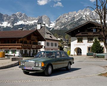 Ein elegantes Auto fährt durch ein malerisches Dorf mit traditionellen Gebäuden. Im Hintergrund sind majestätische Berge und eine klare Himmelsaufnahme zu sehen.