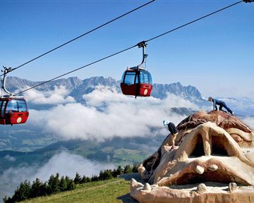 Eine beeindruckende Berglandschaft mit Gondeln, die über Wolken schweben. Im Vordergrund steht eine große Skulptur eines Gesichts.