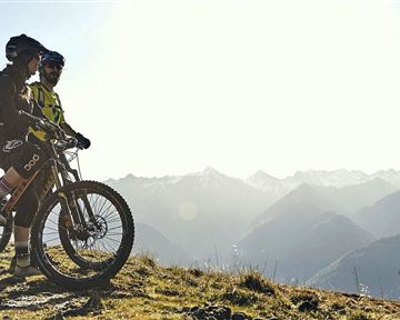 Zwei Radfahrer stehen auf einem Höhenweg und betrachten die Berglandschaft. Die Sonne scheint über den Gipfeln und sorgt für eine beeindruckende Aussicht.
