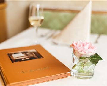 Ein eleganter Tisch mit einer Speisekarte und einer Rose in einer Glasvase. Im Hintergrund steht ein Glas Wein und ein gefaltetes Tuch auf dem Tisch.