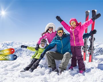 Eine fröhliche Familie steht im Schnee und posiert für ein Foto. Im Hintergrund sind schneebedeckte Berge und Skier sichtbar.