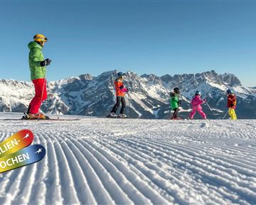 Eine Gruppe von Skifahrern auf einer schneebedeckten Piste mit beeindruckenden Bergen im Hintergrund. Der Himmel ist blau und die Szene zeigt Aktivitäten im Schnee.