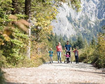 Eine Familie spaziert auf einem malerischen Weg umgeben von Bäumen und Bergen. Die Kinder laufen fröhlich neben den Erwachsenen, während ein Buggy geschoben wird.