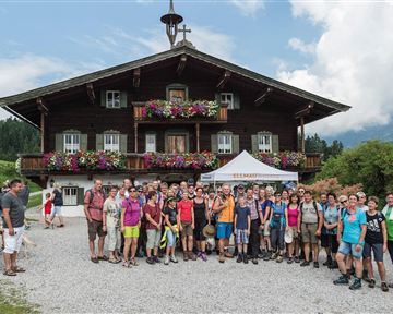Eine große Gruppe von Menschen steht vor einem traditionellen Holzhaus in den Bergen. Das Haus ist mit Blumen geschmückt, und die Landschaft ist grün und bergig.