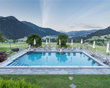 A beautiful pool with sun loungers, surrounded by green meadows and mountains. The sky is clear and shows soft colors of the sunset.
