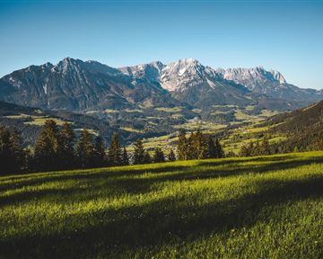 A green meadow with views of impressive mountain peaks and a bright blue sky. The landscape is calm and naturally beautiful.
