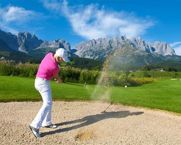 Ein Golfer schlägt aus einem Bunker auf einem schönen Golfplatz mit beeindruckenden Bergen im Hintergrund. Der Himmel ist klar und blau, die Landschaft ist grün und einladend.