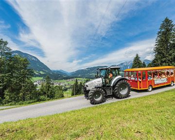 A tractor is pulling a colorful wagon with passengers on a winding road. In the background, green hills and a clear sky can be seen.