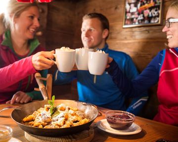 Drei Freunde stoßen mit Tassen an und genießen eine Mahlzeit in einer gemütlichen Umgebung. Auf dem Tisch liegt ein Teller mit köstlichen Leckereien.