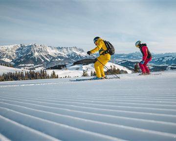 Zwei Skifahrer fahren über eine präparierte Piste in den Bergen. Im Hintergrund sind schneebedeckte Berge und ein blauer Himmel zu sehen.