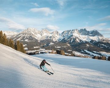A skier is sliding down a snow-covered slope. In the background, majestic mountains and a clear sky can be seen.