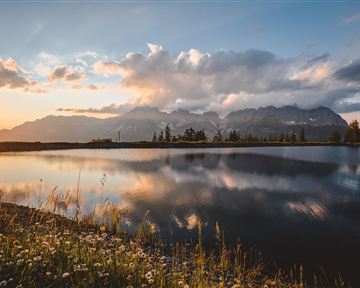 A picturesque lake surrounded by mountains and blooming meadows. The setting sun reflects gently on the water.