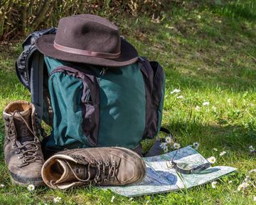 A backpack is standing on a meadow, next to a pair of hiking boots and a map. A hat lies on the backpack and in the background, fresh grass and small flowers can be seen.