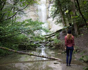 A woman stands at a small waterfall in a forested area. The forest is green and lush, with a clear water pool in the foreground.