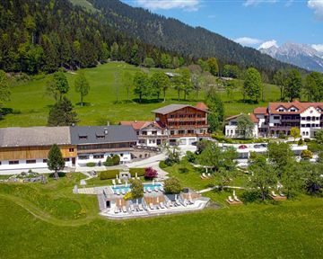 A picturesque landscape with a hotel complex, surrounded by forests and mountains. In the foreground, there is a pool area and green meadows.