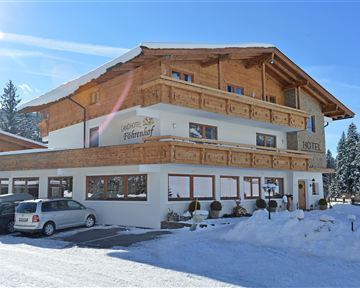 A charming hotel in winter with wooden cladding and a snow-covered ground. The surroundings are adorned with trees and the sky is clear and blue.