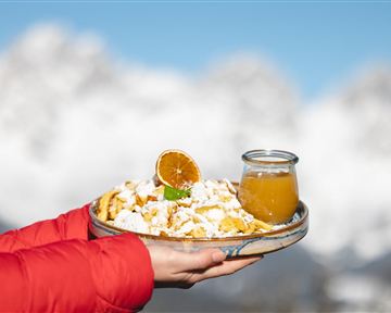 A plate of freshly made Kaiserschmarrn, decorated with powdered sugar and a dried orange slice. Next to it is a glass of fruit juice, set against an impressive mountain backdrop.