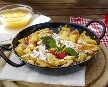 Ein köstlicher Kaiserschmarrn in einer schwarzen Pfanne, garniert mit Puderzucker und einer Frischeminze. Daneben befindet sich eine Schüssel mit Apfelmus.