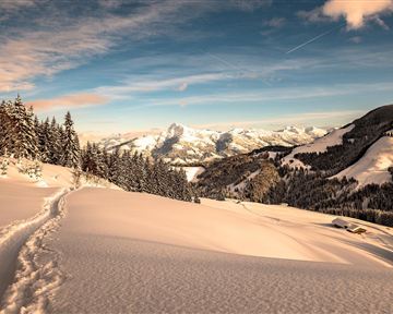 A snowy landscape with gentle hills and high mountains in the background. The sky is clear and bright blue.