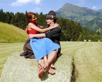 Ein Paar sitzt auf einer Heuballen in einer wunderschönen, grünen Landschaft. Die Umgebung ist hügelig, mit Bäumen und einem klaren Himmel im Hintergrund.