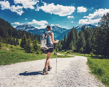 A person is hiking on a gravel path through a green landscape. In the background, there are mountains and a blue sky with some clouds.