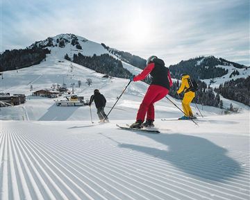 Three skiers enjoy the slopes in a wintry landscape. The sun shines over the snow-covered mountains.