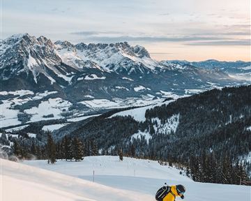 A skier in a yellow suit is skiing down the snowy slope. In the background, majestic mountains and a serene winter landscape can be seen.