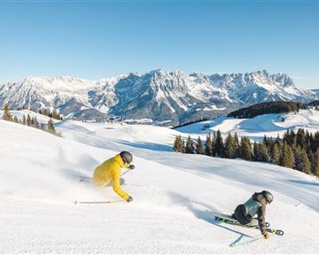 Two skiers are skiing down a snow-covered slope in the mountains. The sky is clear and the landscape is picturesque.