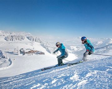 Two skiers glide down the snowy slope. In the background, snow-covered mountains stretch out under a clear blue sky.