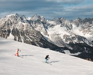 Zwei Skifahrer fahren die verschneite Piste hinunter. Im Hintergrund sind imposante Berge unter einem grauen Himmel zu sehen.