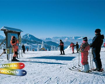 Eine Gruppe von Skifahrern steht auf einer schneebedeckten Piste mit beeindruckenden Bergen im Hintergrund. Die Szene zeigt fröhliche Familien, die sich auf die Skiaktion vorbereiten.