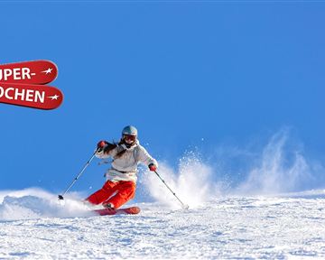 Ein Skifahrer fährt über frischen Schnee unter einem klaren blauen Himmel. Im Hintergrund sind Schilder mit der Aufschrift "SUPER-SKIWOCHEN" zu sehen.