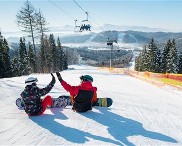 Zwei Snowboarder sitzen im Schnee und feiern gemeinsam. Im Hintergrund sind schneebedeckte Berge und eine Seilbahn zu sehen.