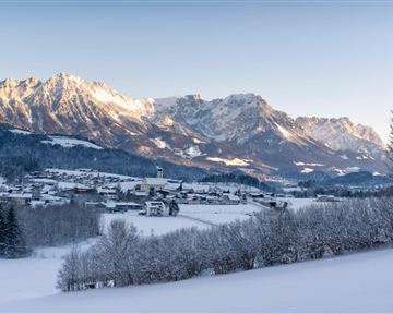 A winter landscape with snow-covered mountains and a small village in the valley. The landscape is illuminated by gentle rays of light from the low-hanging sun.