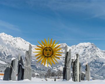 A winter landscape with snow-covered mountains and an artistic sun sculpture in the center. The scene radiates a peaceful and impressive atmosphere.
