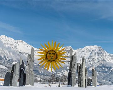 A winter landscape with snow-covered mountains and a colorful sun symbol in the center. Large stones surround the symbol, creating a mystical atmosphere.