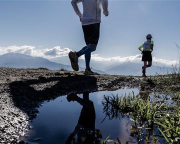 Zwei Läufer joggen auf einem unebenen Weg mit Blick auf die Berge. Im Vordergrund spiegelt sich der Himmel in einer Pfütze.