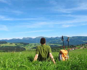 Ein Wanderer sitzt auf einer grünen Wiese und schaut auf die Berge. Der Himmel ist klar und blau, im Hintergrund sind sanfte Hügel und ein kleines Dorf zu sehen.