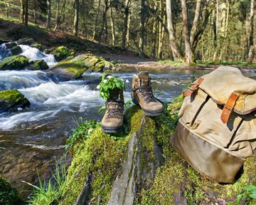 Wanderstiefel und ein Rucksack stehen an einem Bachlauf in einem Wald. Die Umgebung ist grün und ruhigen, ideal für Outdoor-Abenteuer.