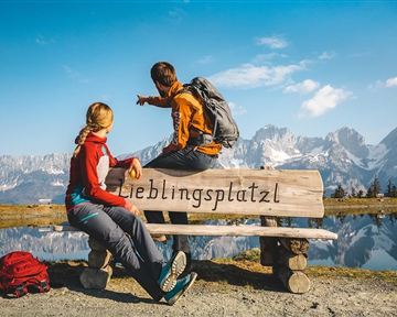 Zwei Wanderer sitzen auf einer Bank mit der Aufschrift „Lieblingsplatzl“ und blicken auf die beeindruckenden Berge. Die Landschaft ist klar und sonnig, umgeben von Wasser und Natur.