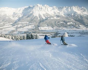 Zwei Skifahrer fahren über eine verschneite Piste in den Bergen. Im Hintergrund sind majestätische Schneegipfel und eine klare Winterlandschaft sichtbar.