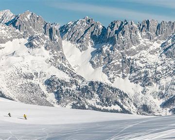 Eine verschneite Landschaft mit majestätischen Bergen im Hintergrund. Zwei Skifahrer gleiten über den glitzernden Schnee.
