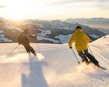 Two skiers are riding in the evening sun over a snow-covered slope. The mountains in the background provide a picturesque backdrop.